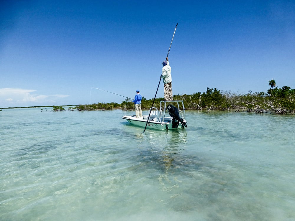 Guide quietly poles an angler aboard his skiff across a shallow flat.